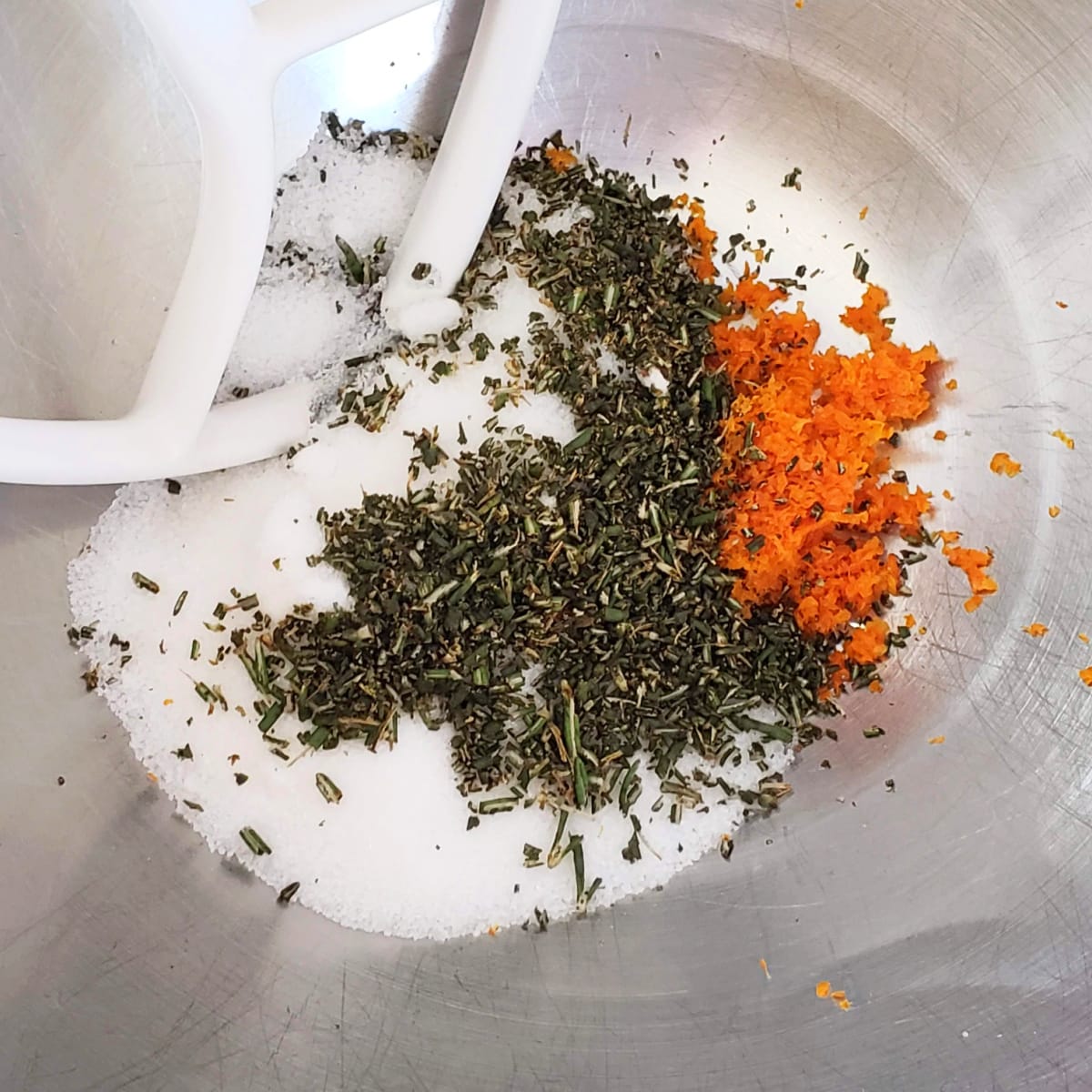 Sugar, rosemary and orange zest in the bottom of a silver mixing bowl, with white mixer paddle