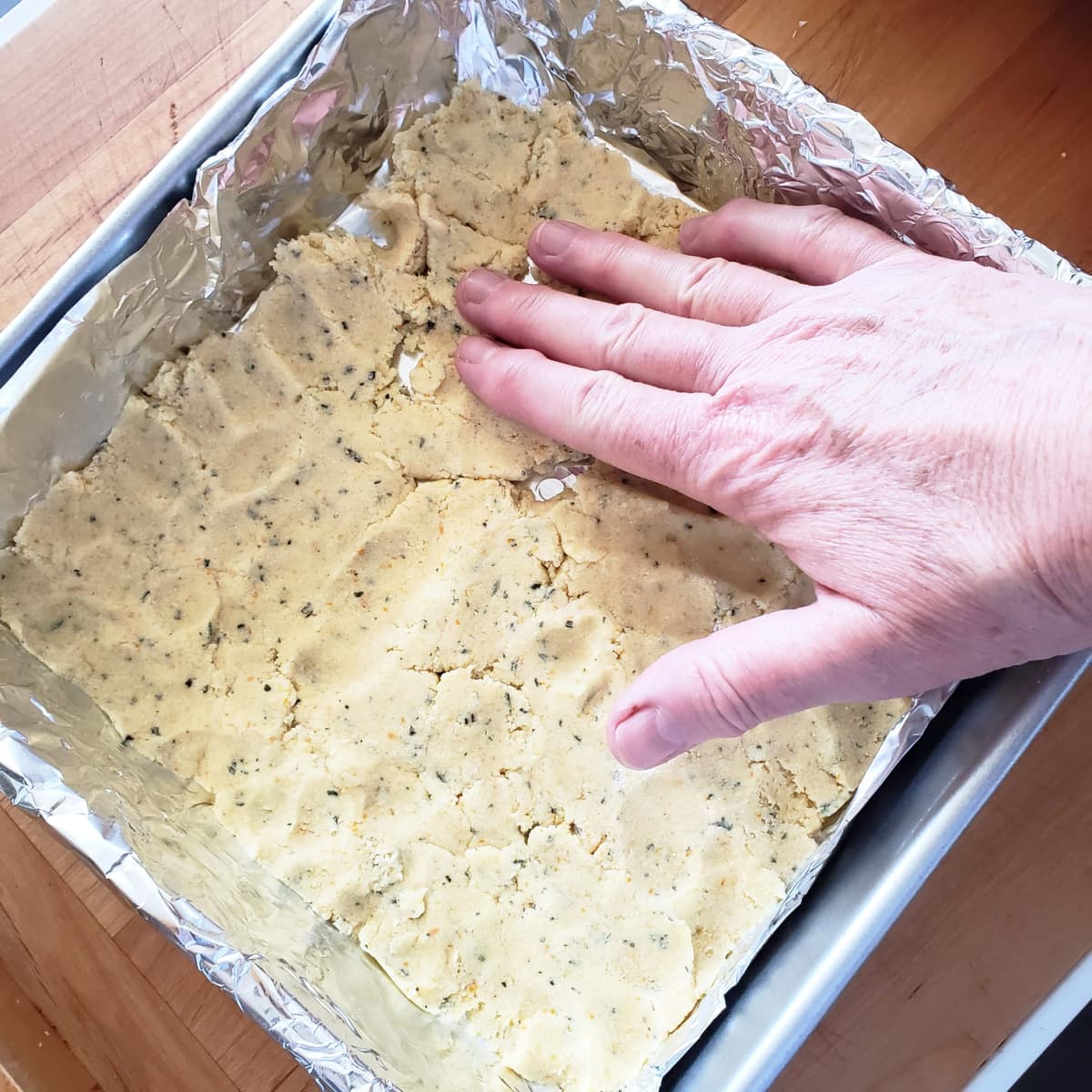 A hand with outstretched fingers pats dough into a silver foil-lined baking pan