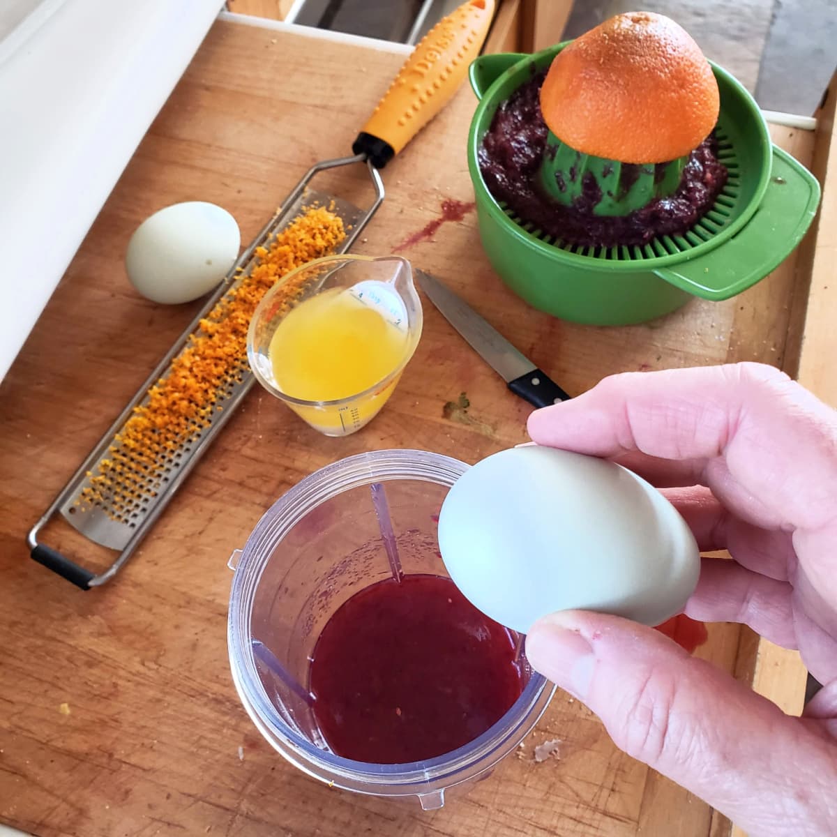 Array of ingredients on a wooden cutting board, with a hand holding a light blue egg over the edge of a container ready to crack it