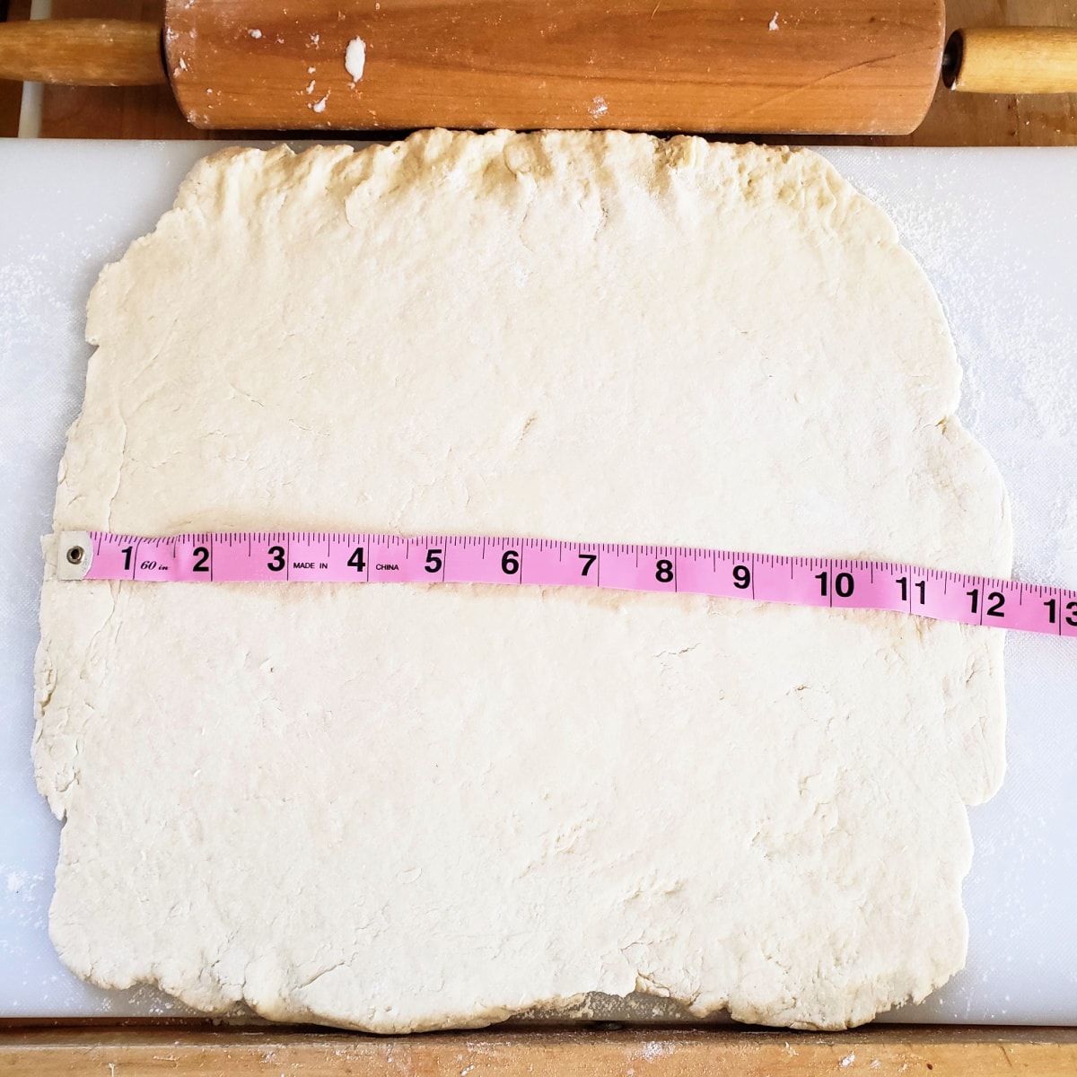 Dough rolled out square with a pink tape measure across it and a rolling pin above it