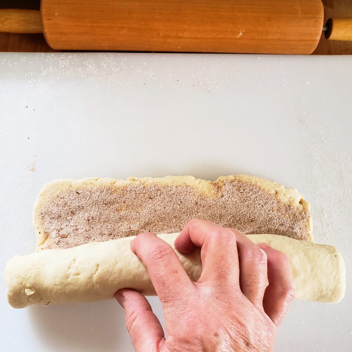 A hand rolls dough into a long log on a white cutting board with rolling pin at the top