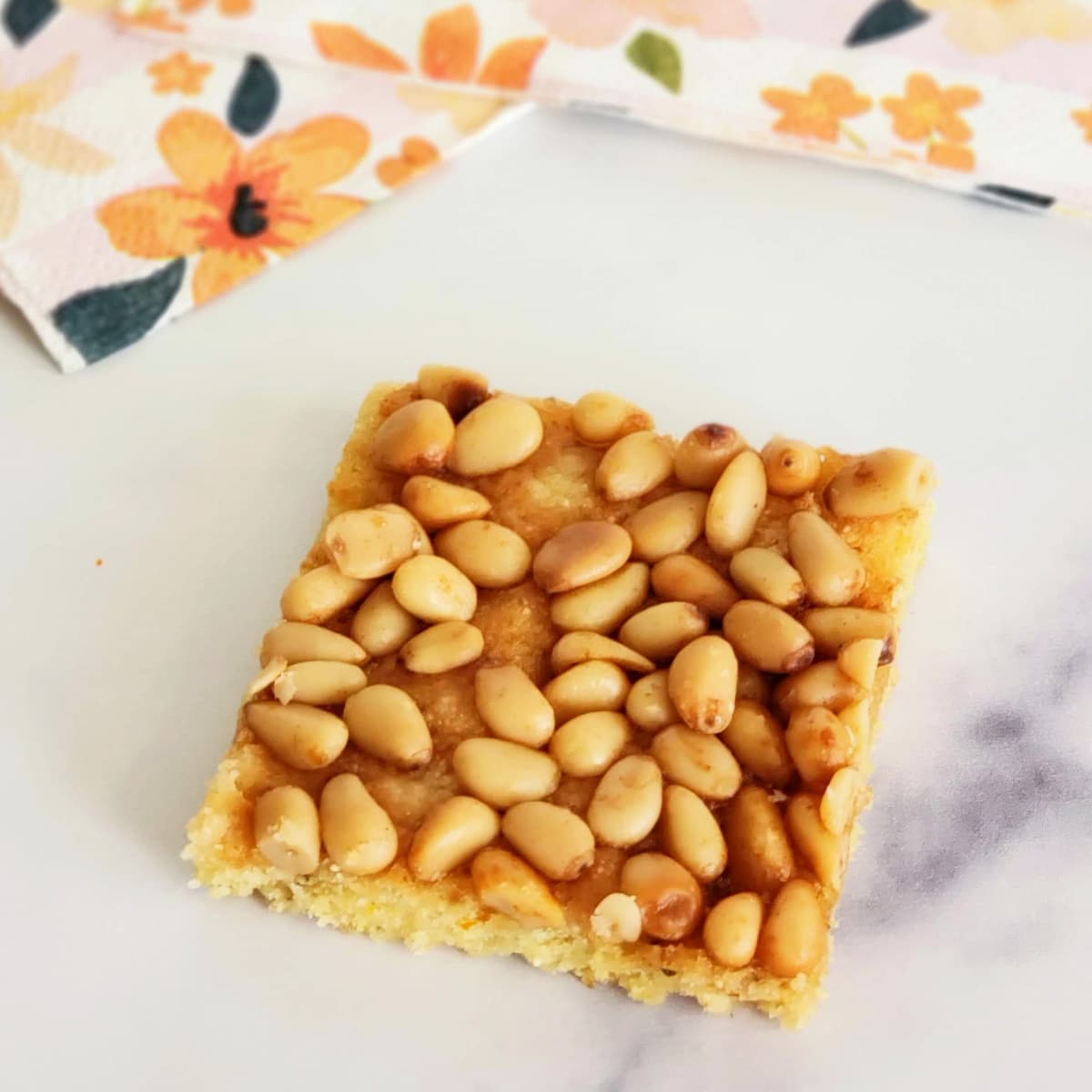 Square cookie covered in pine nuts on a white marble countertop with flowered napkins behind