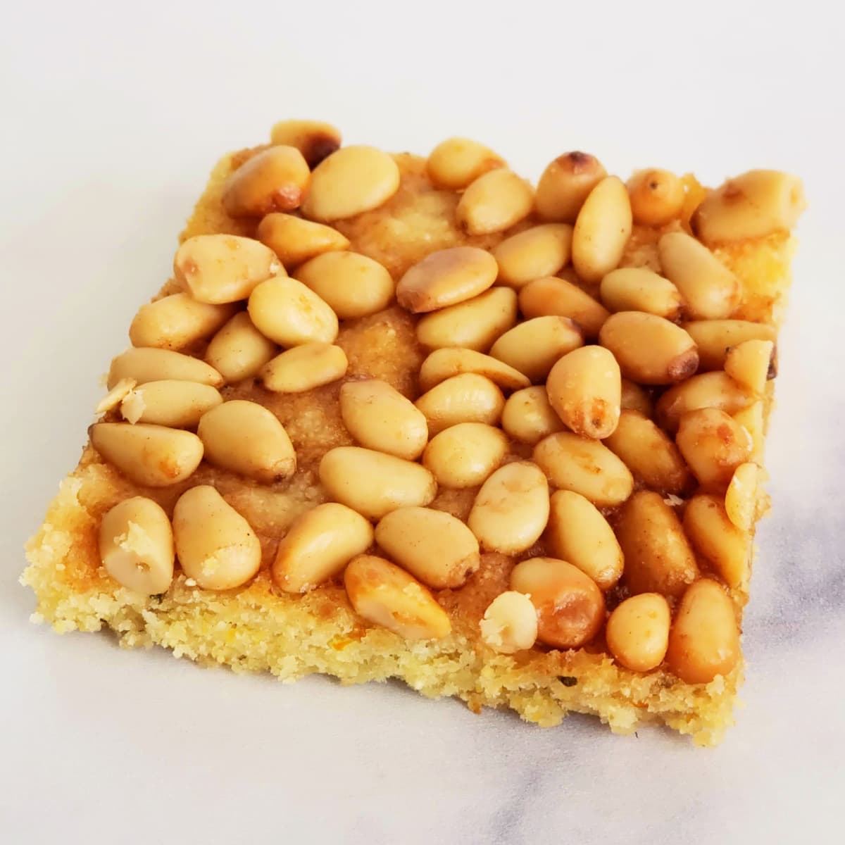 Square bar cookie Rosemary Pine Nut Cornmeal Shortbread rests on a white marble countertop
