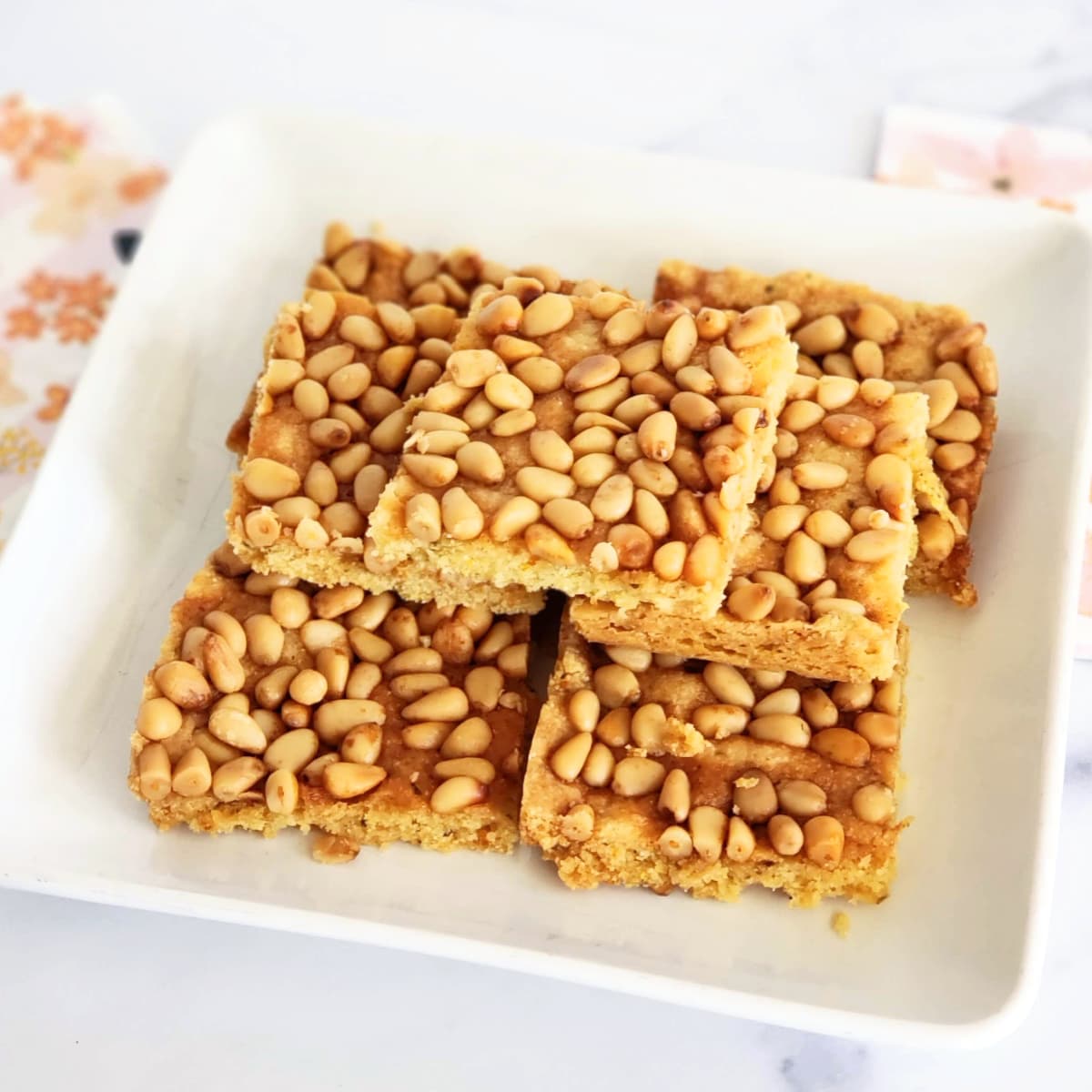 7 pine nut cookies stacked on a square white plate atop flowered napkins