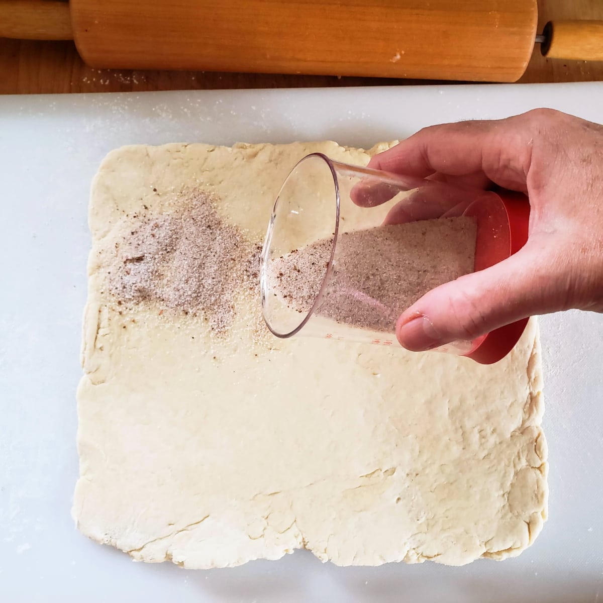 A hand holds a clear measuring cup and sprinkles cinnamon sugar on square piece of dough