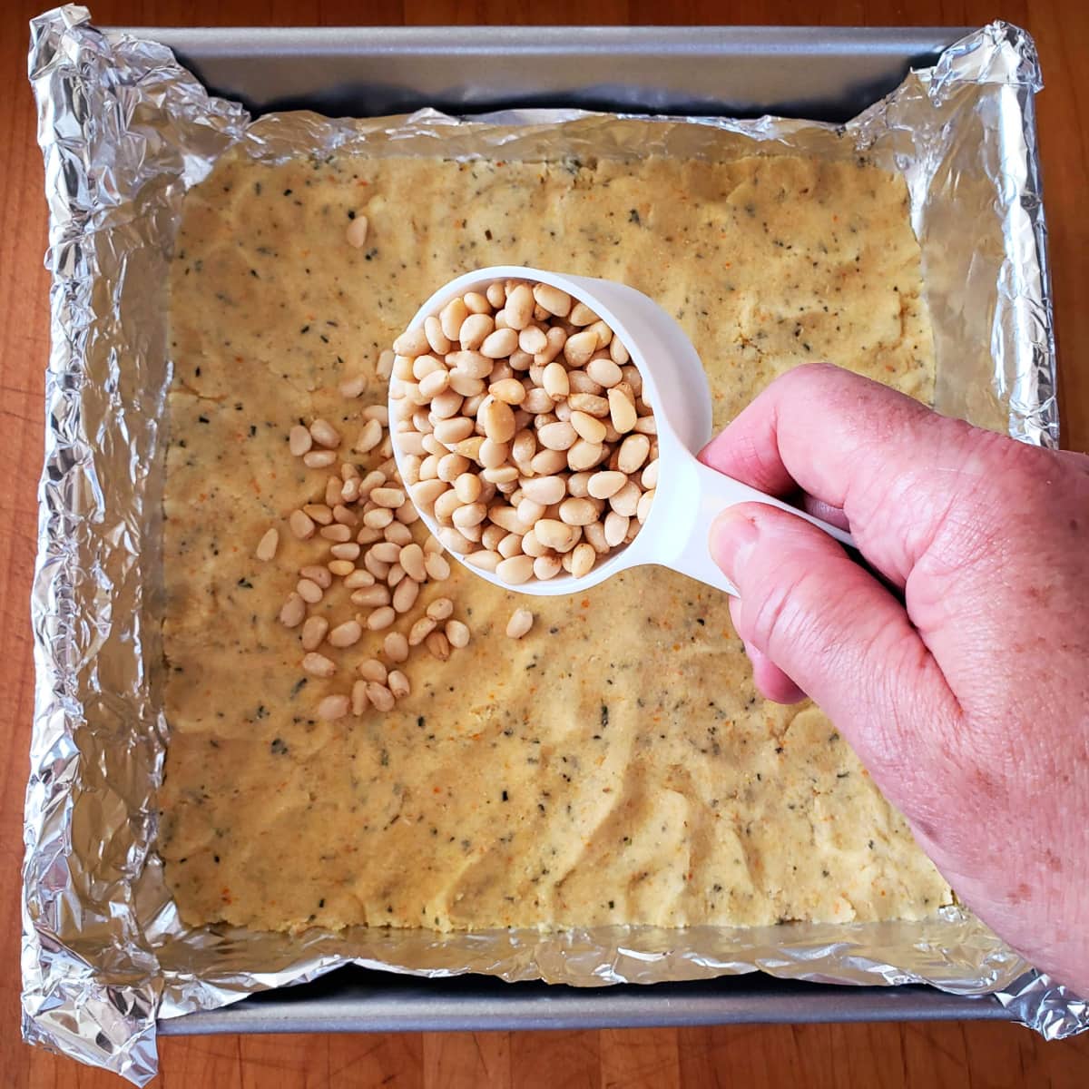 Hand holding a white measuring cup filled with pine nuts sprinkles the nuts on top of dough in silver baking dish