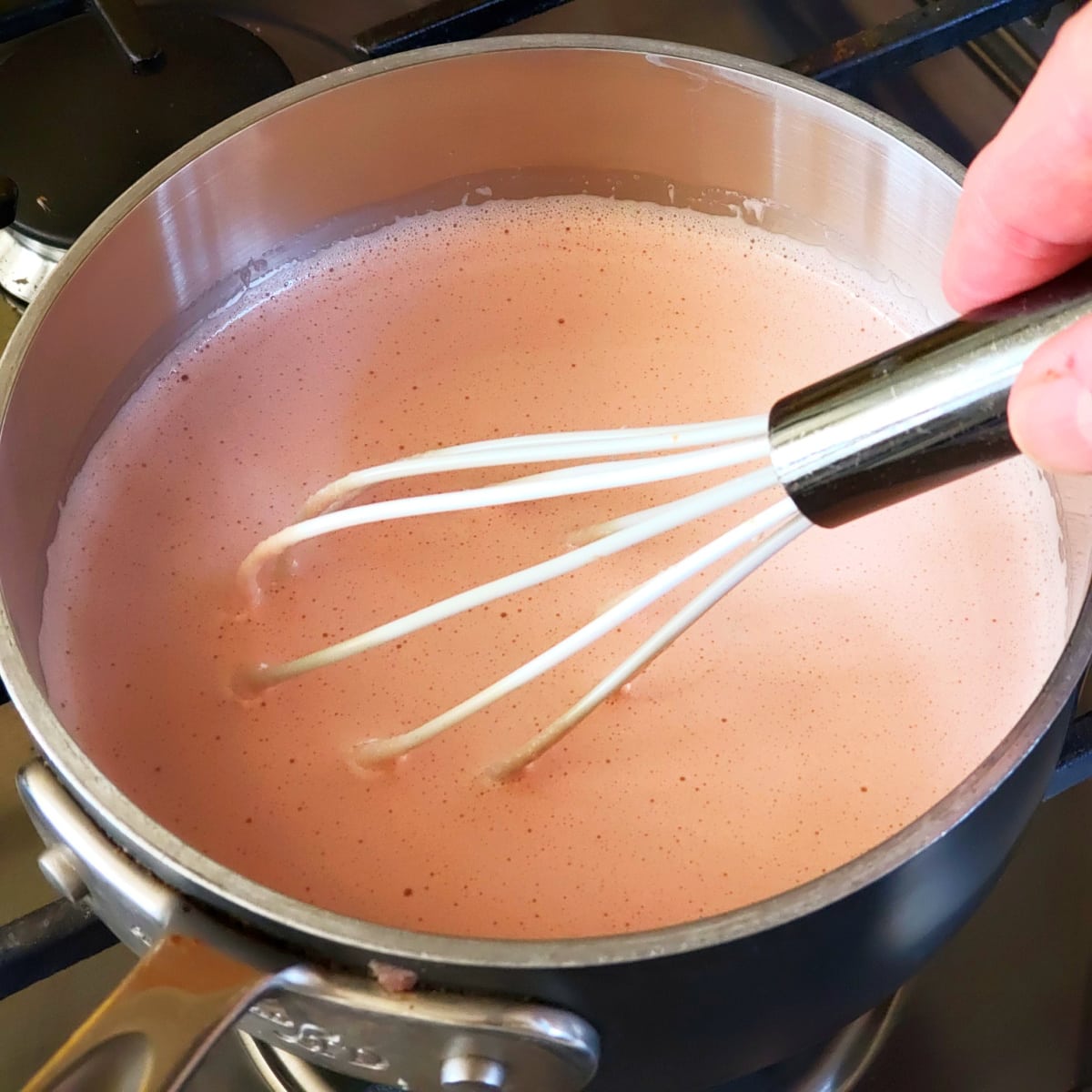 A hand holds a white whisk to stir orange curd in a silver saucepan