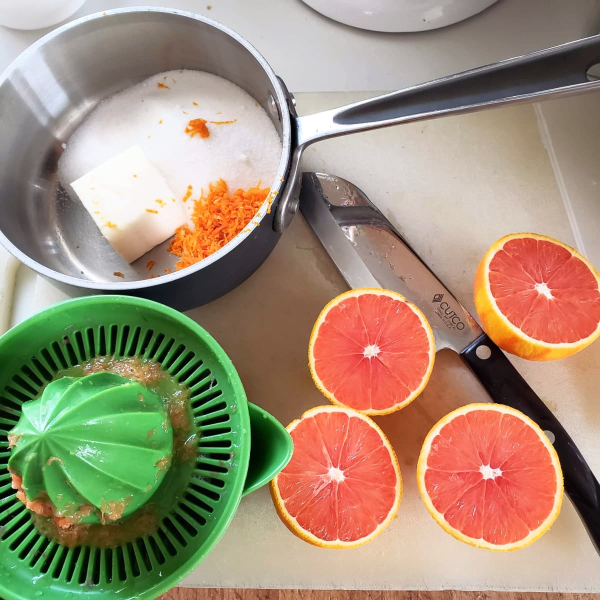 4 orange halves sit on a white cutting board next to a green juicer, knives and a small saucepan