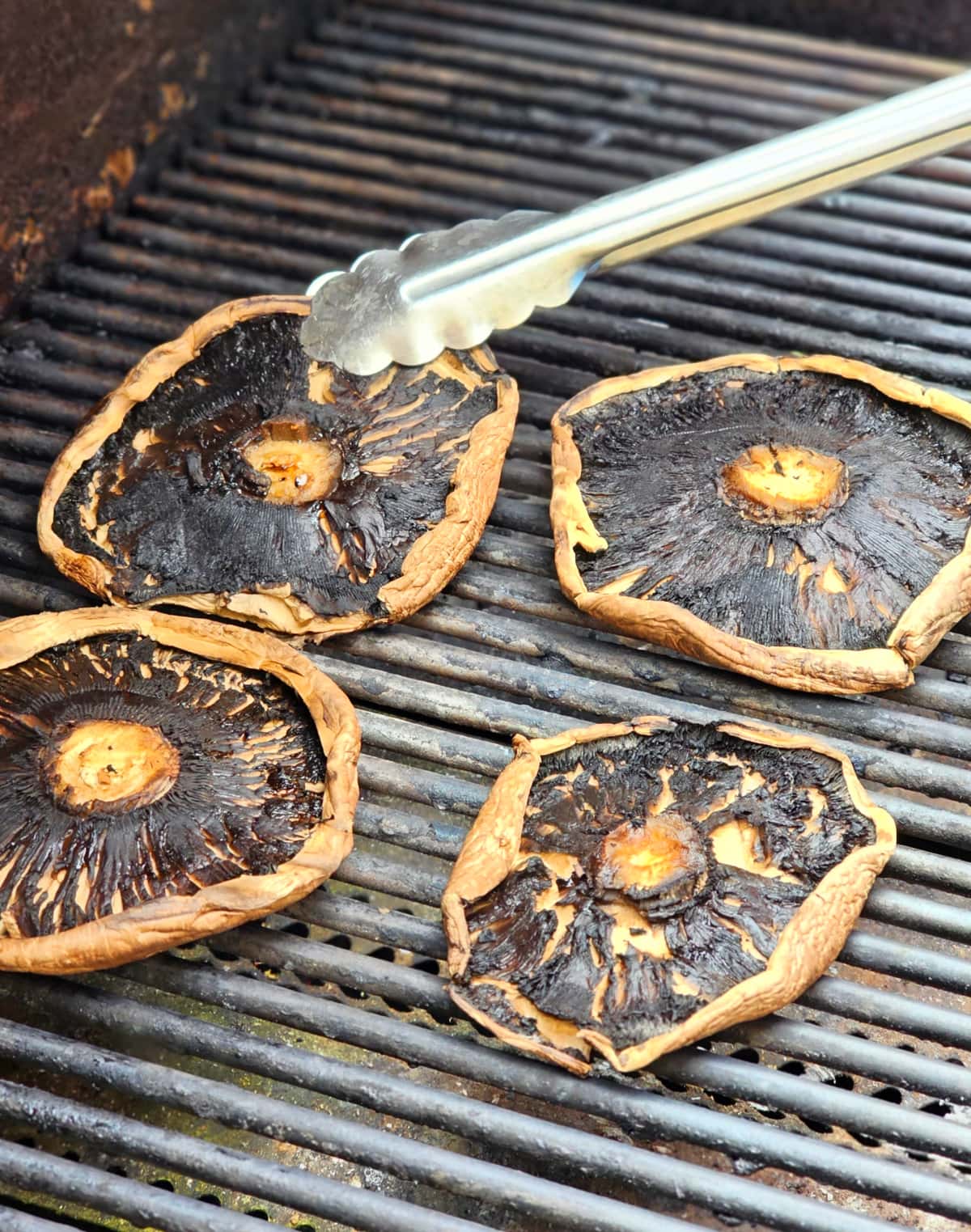 4 Portabella mushrooms on the grill with a metal tongs holding the edge of one. 