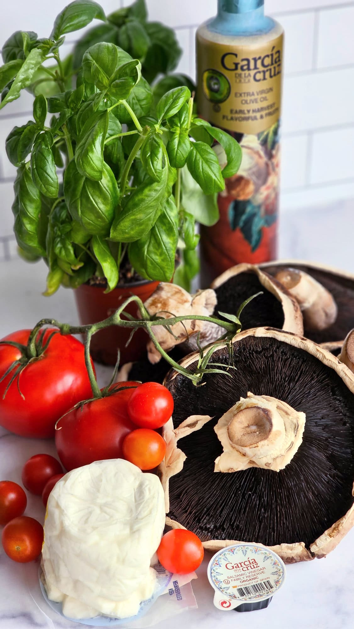 Ingredients for a Grilled Portabella Caprese including tomatoes, mushrooms, basil, and olive oil on a white marble counter. 