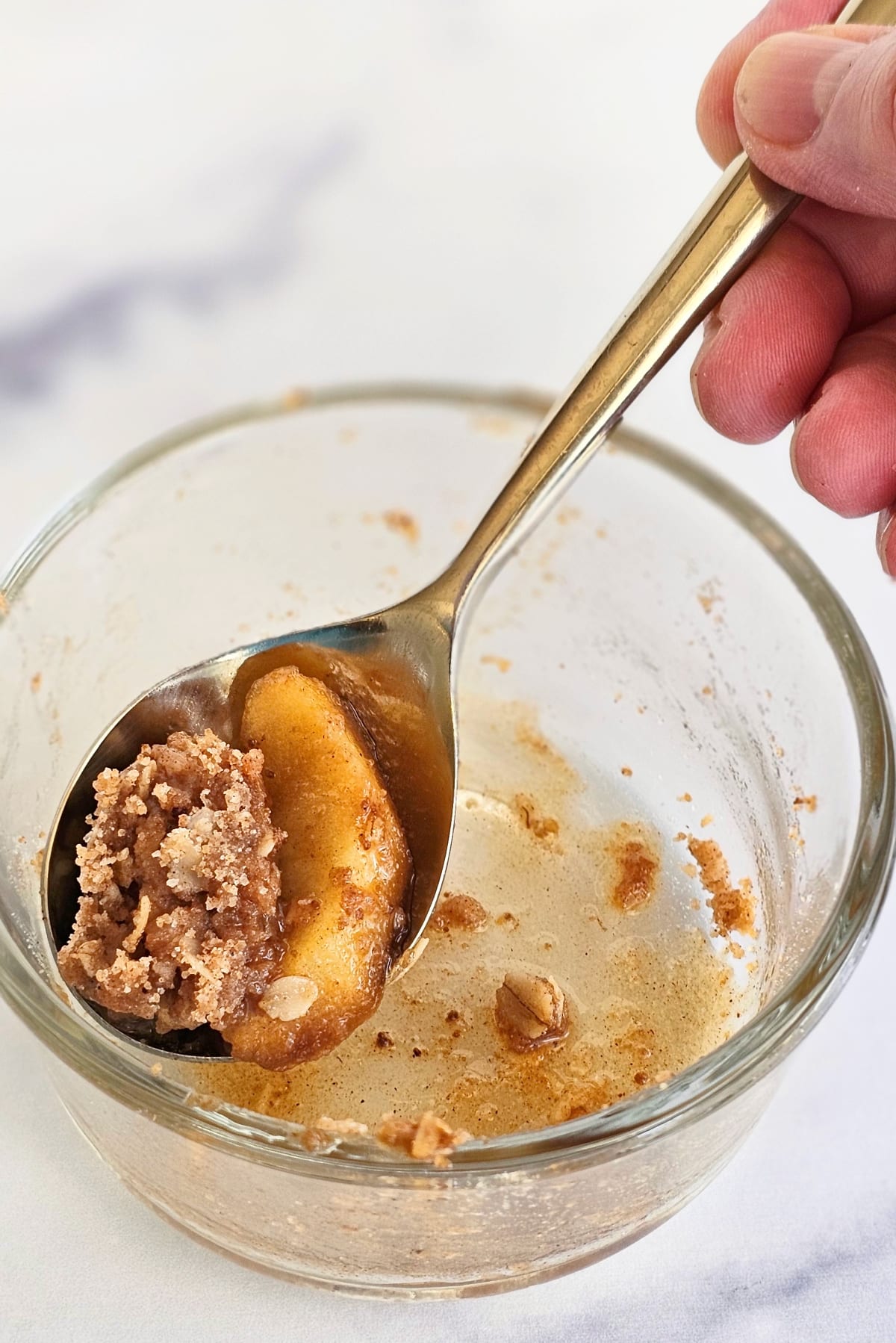 Hand holds a spoon containing the last bit of apple crisp scooped from a glass bowl on a white marble countertop
