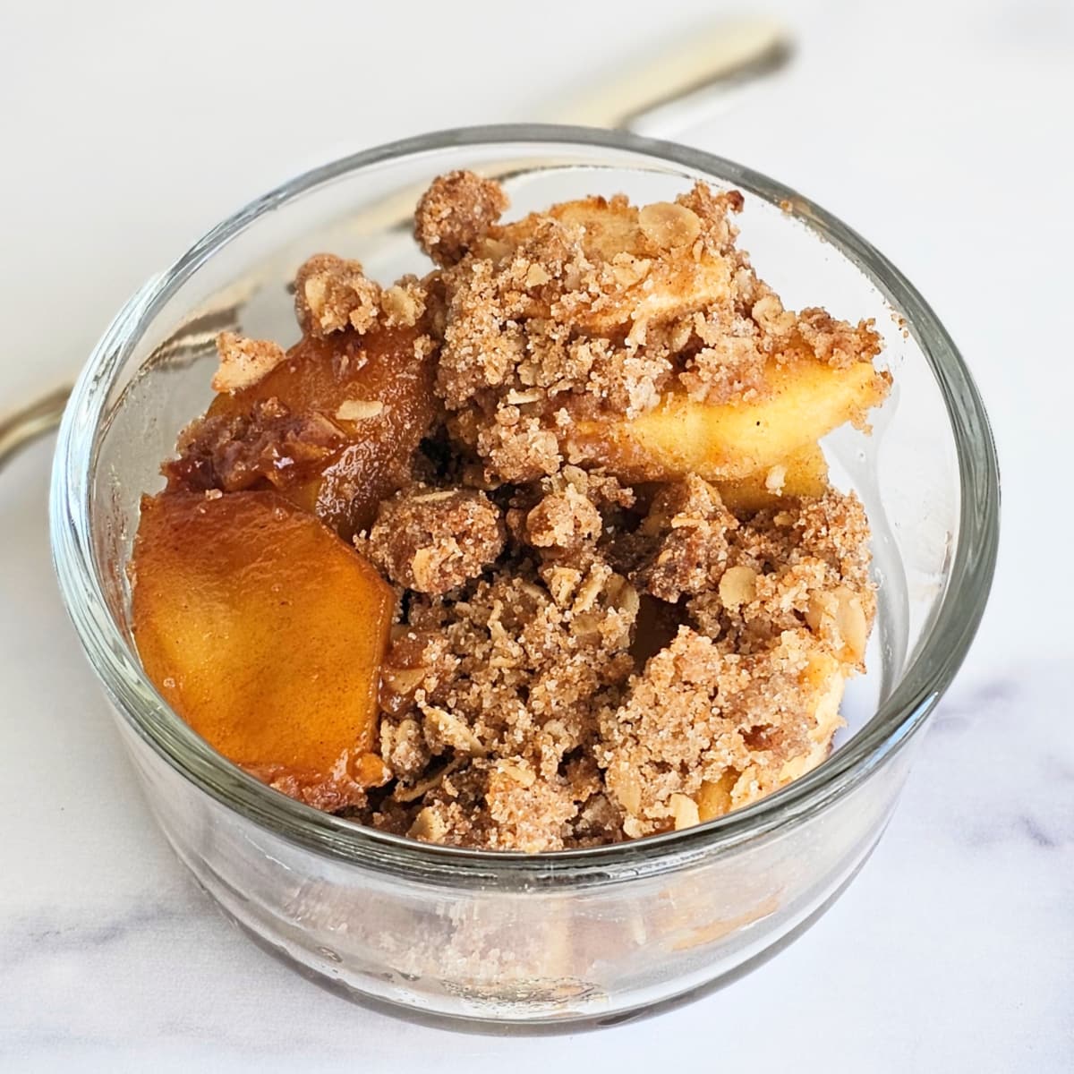 Apple Crisp with Streusel Topping in a glass bowl with a spoon along the back on a white marble counter top
