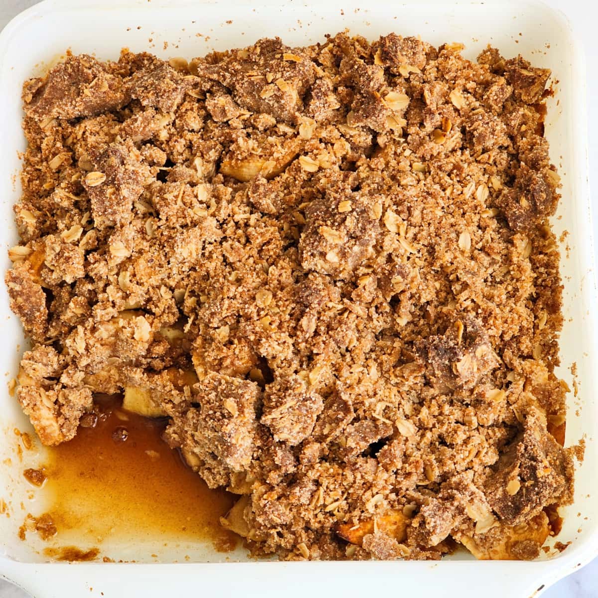 White square baking dish with Apple Crisp with Streusel Topping with bottom left piece missing and brown juice left behind