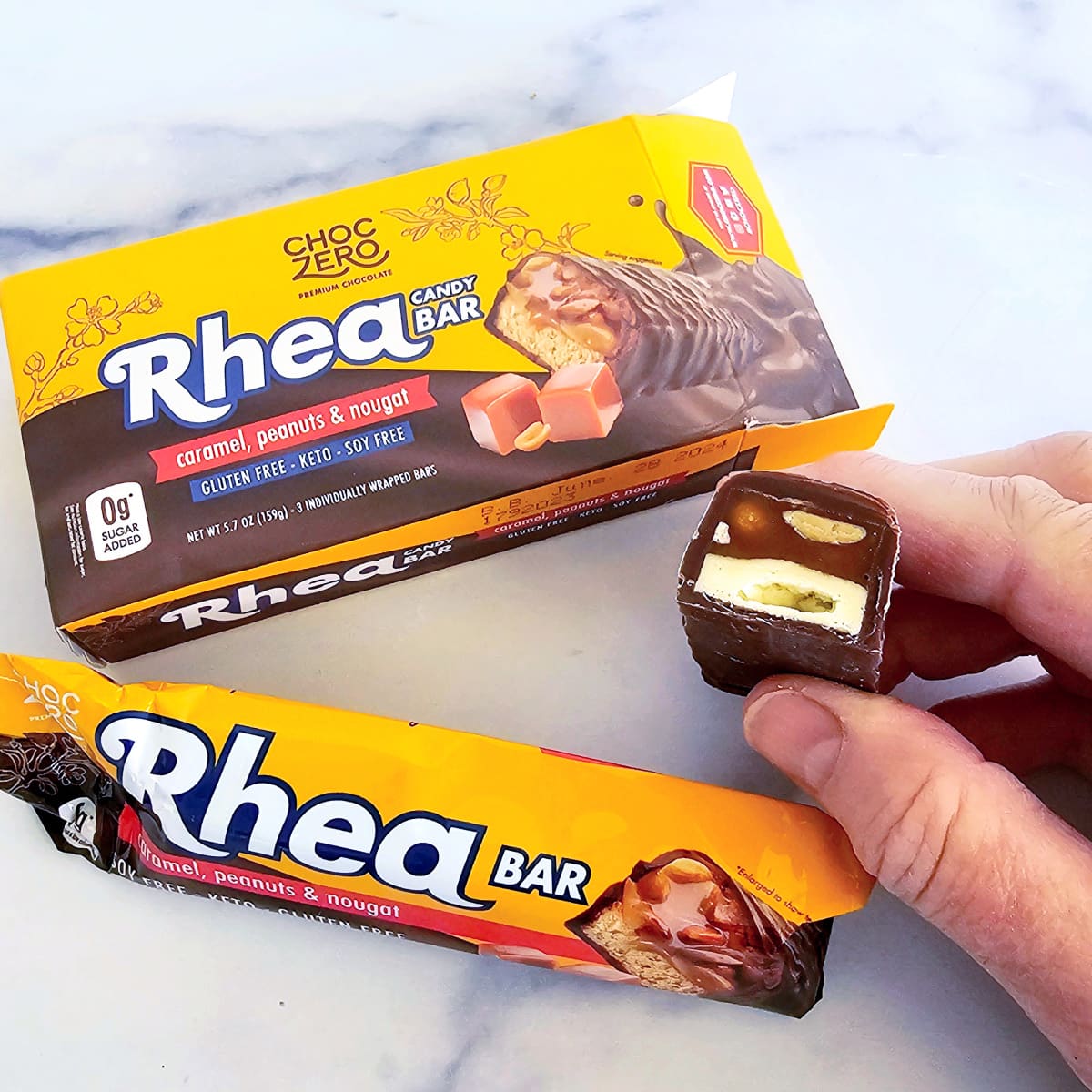 Hand holds a cut Rhea candy bar with a wrapper in front and a box behind