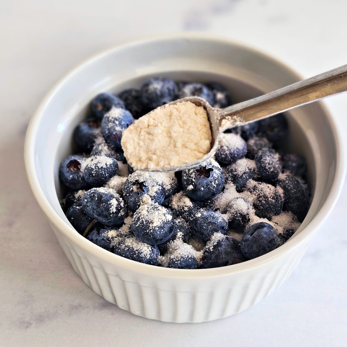 Blueberries in a white ramekin are dusted with sugar while a measuring spoon with white flour rests atop berries