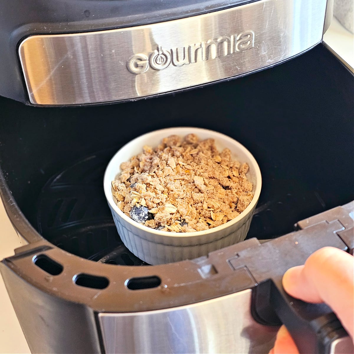 Blueberry crisp in white ramekin in the air fryer, with a hand holding the handle