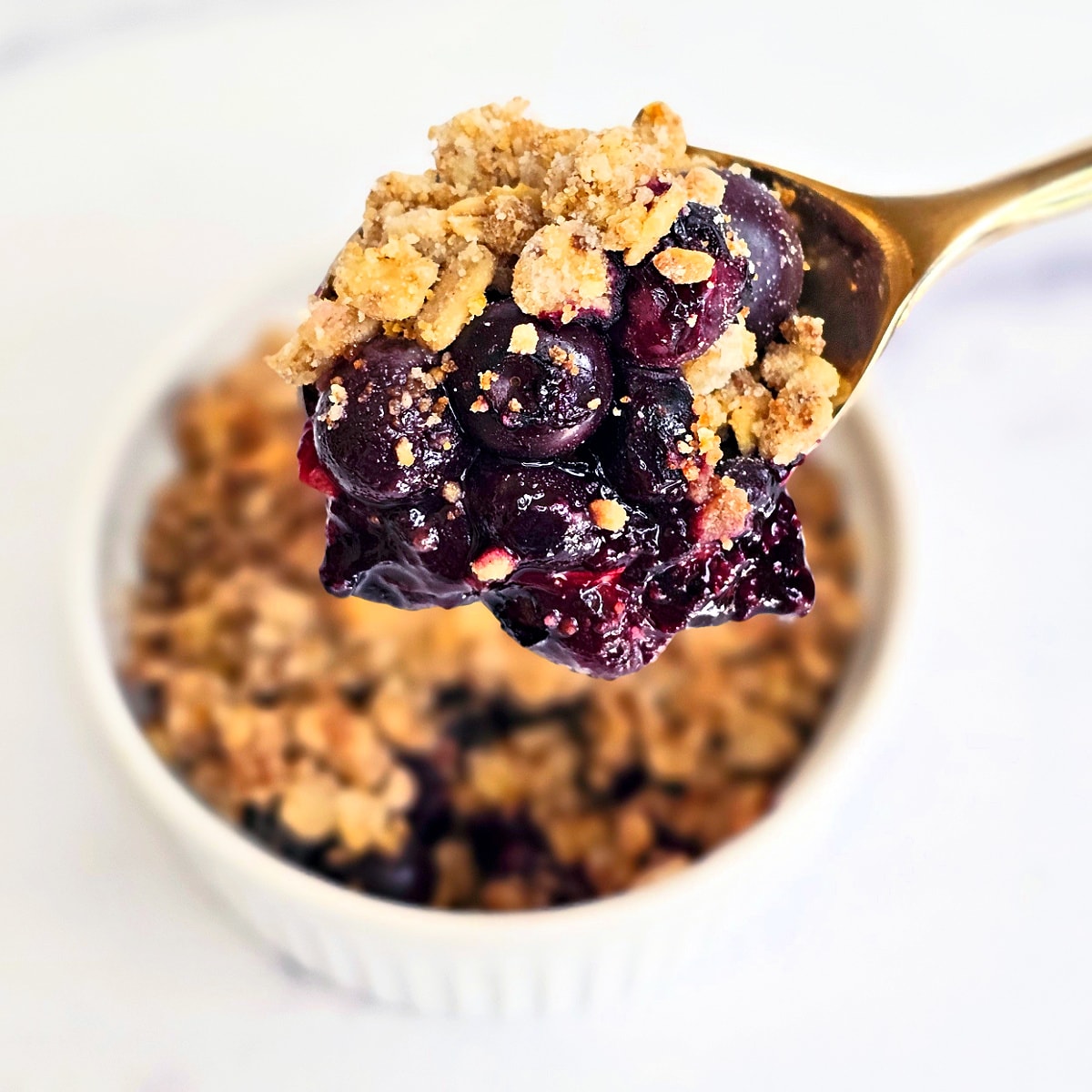 Spoon holds a scoop of Air Fryer Blueberry Crisp above a white ramekin full of the dessert