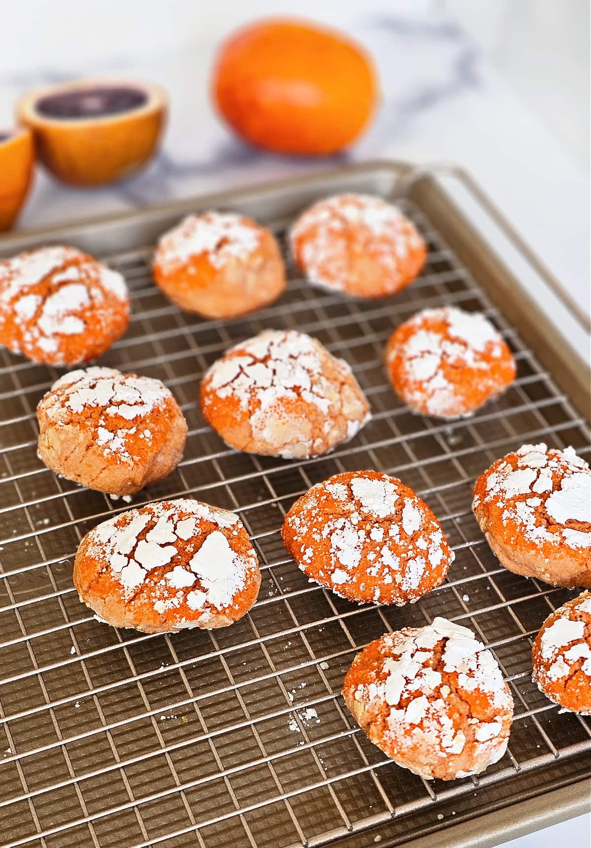 11 orange cookies on a gold colored cooling rack with a whole orange and a cut blood orange in the background out of focus. 