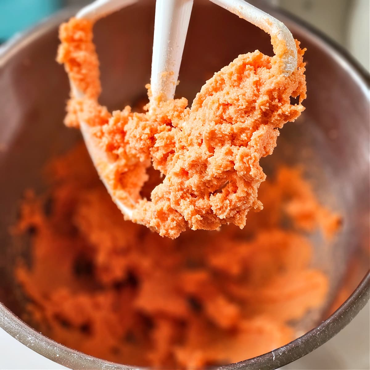 White mixer paddle with bright orange cookie dough on it, with more in the stainless steel bowl below