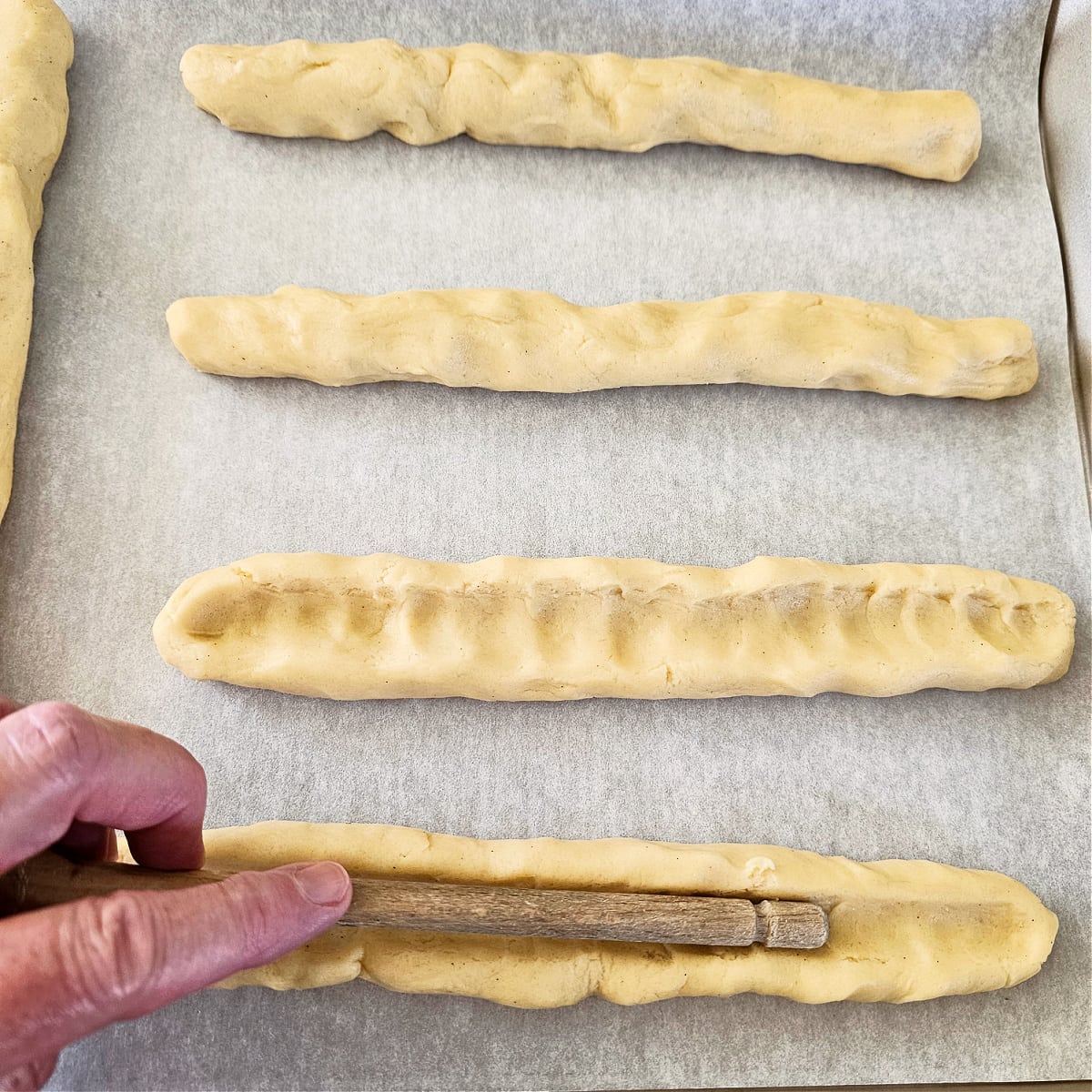 Hand pushes the handle of a wooden spoon into dough log to form trough