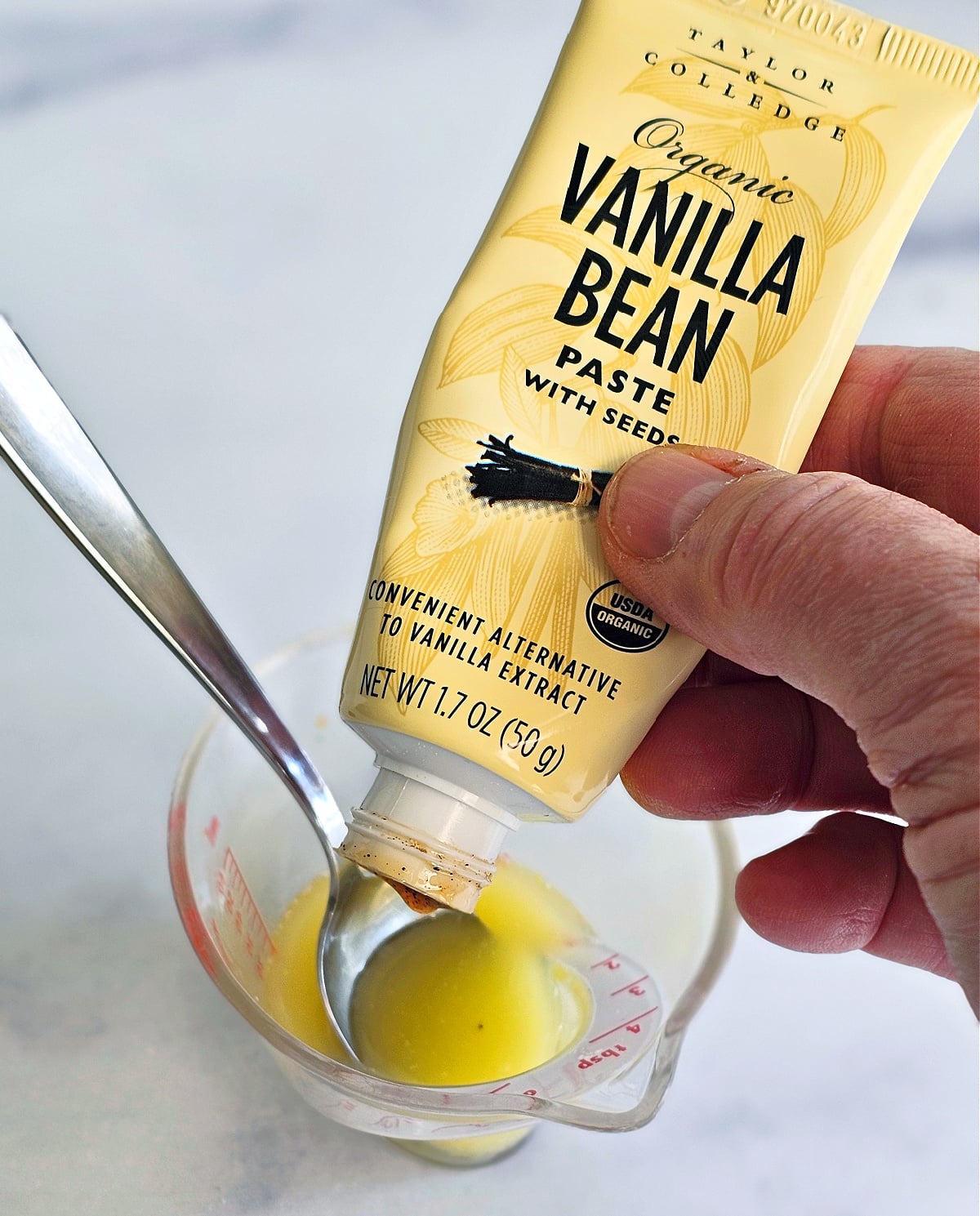 Hand holds tube of vanilla bean paste squirting into cup of melted butter, with a spoon to the left