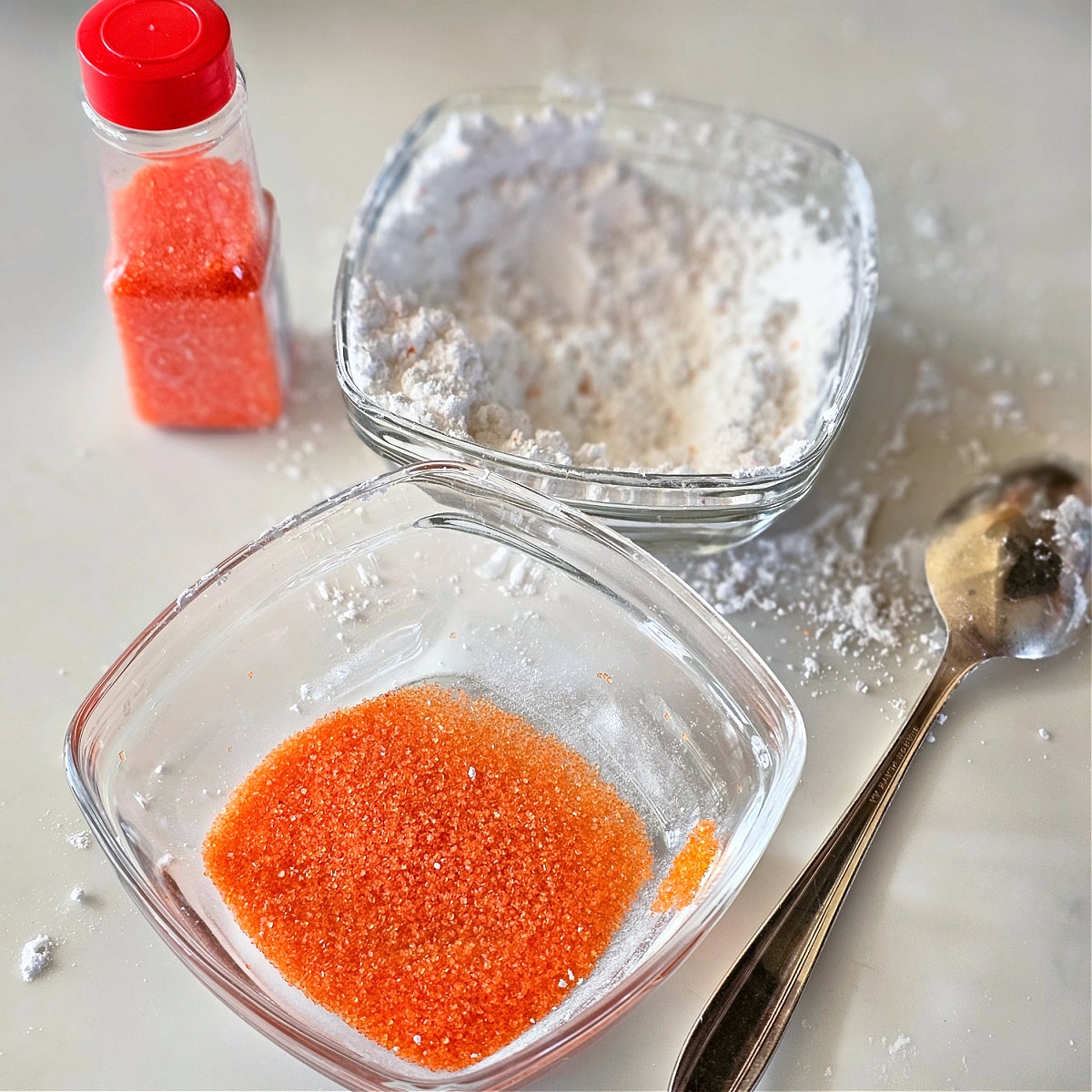 2 square glass bowls, one with powdered sugar, one with orange sanding sugar, with a spoon on the right and a bottle of orange sugar on the right