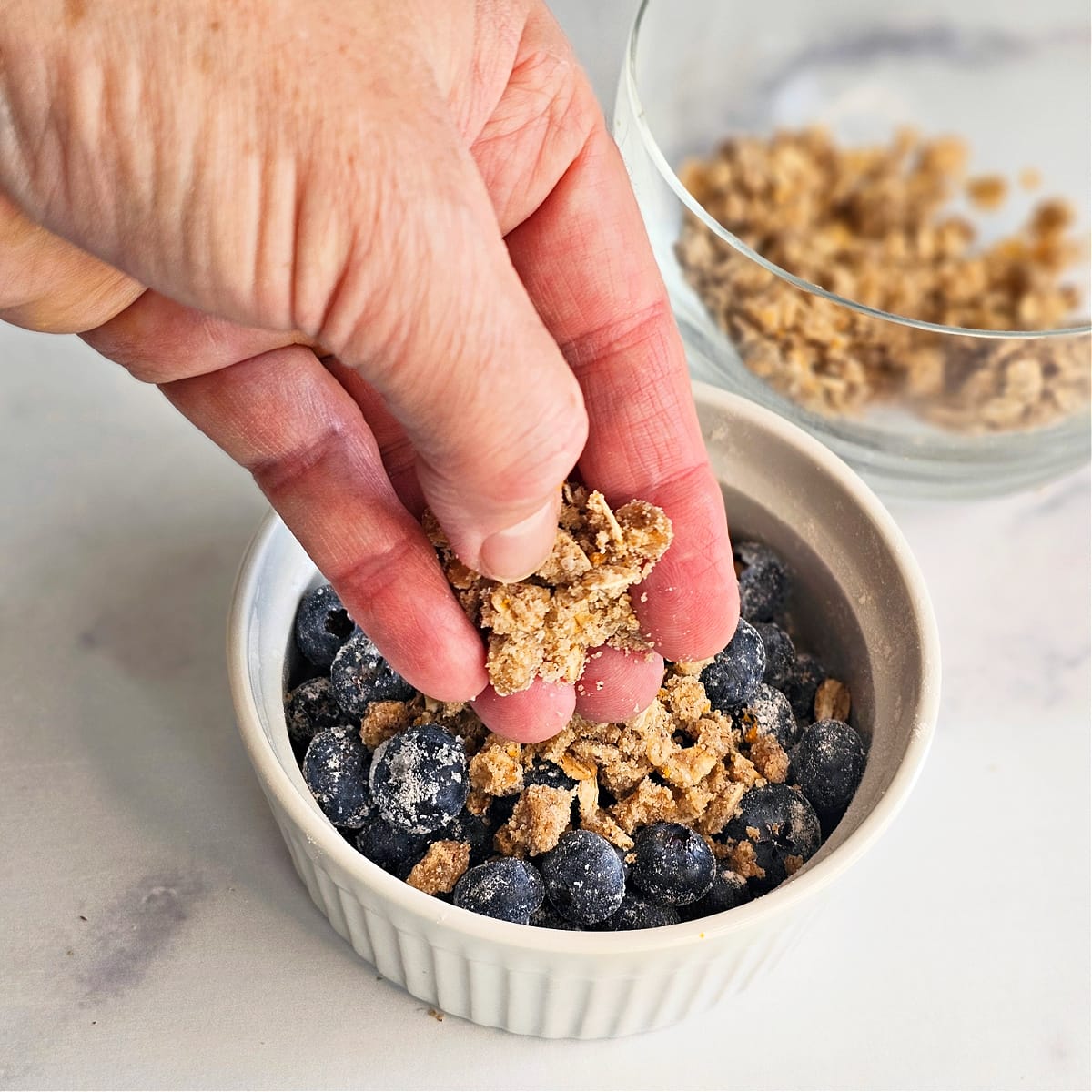 Hand sprinkles topping on Air Fryer Blueberry Crisp in a white ramekin