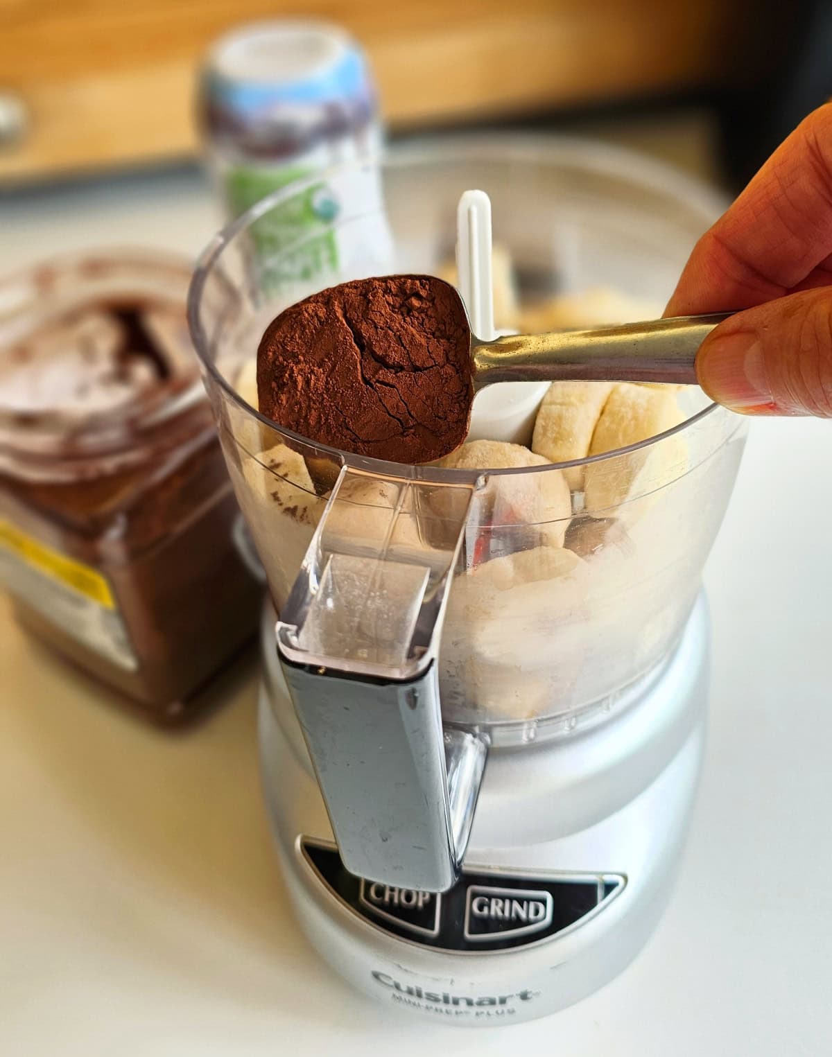 Hand holding a measuring spoon filled with cocoa hovers over a silver mini food processor filled with banana slides, with ingredients in the background