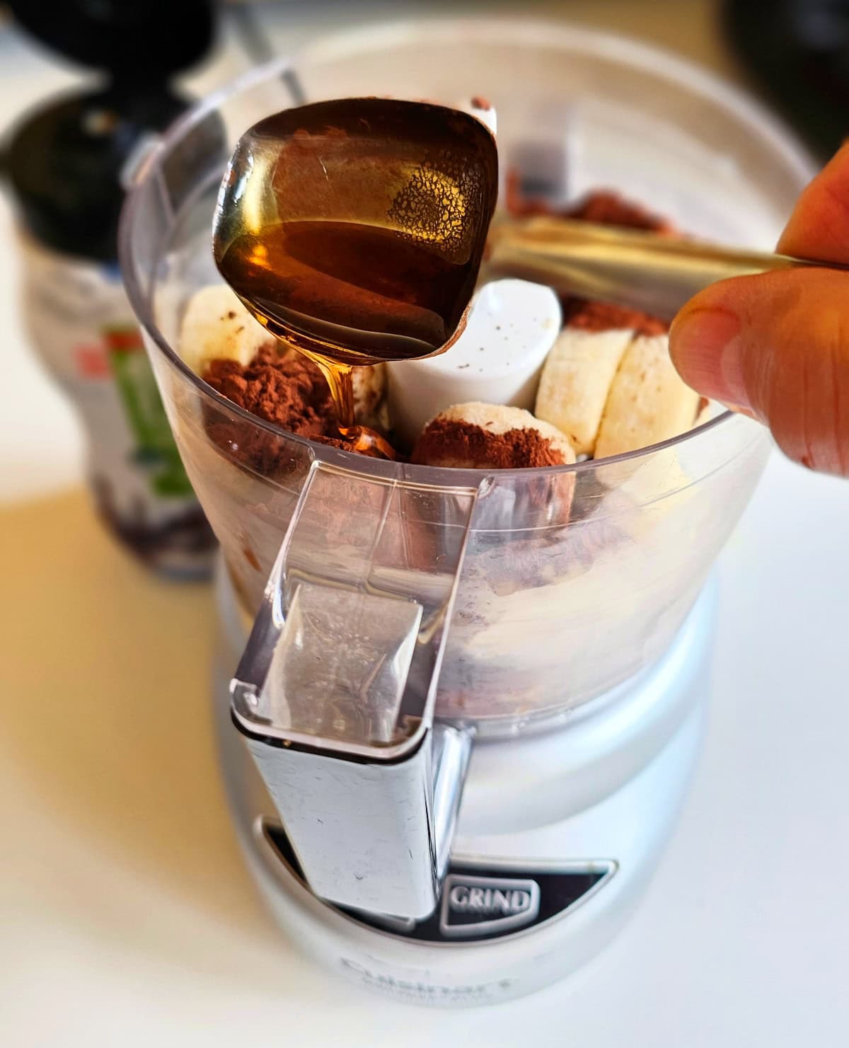 Hand holding a measuring spoon pours maple syrup from that spoon into a mini food processor filled with sliced bananas