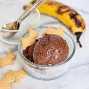 A glass bowl of brown chocolate ice cream is surrounded by cookies and a ripe banana on a white counter