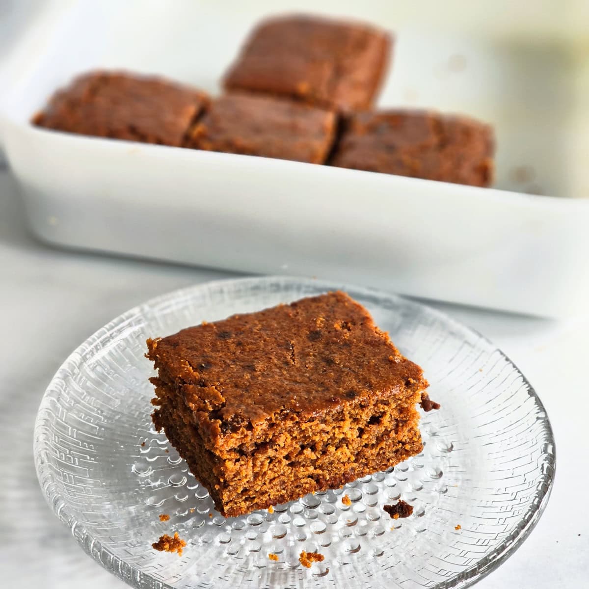 Square piece of brown Molasses Cornbread sits on a glass plate in front of a white baking dish with more cornbread square pieces inside. 