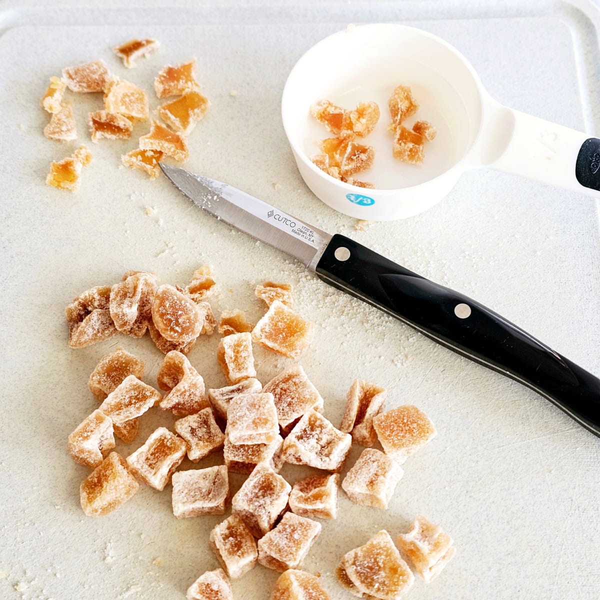 Pieces of candied ginger on a light gray cutting board with a black-handled paring knife and a white measuring cup on the board as well. 