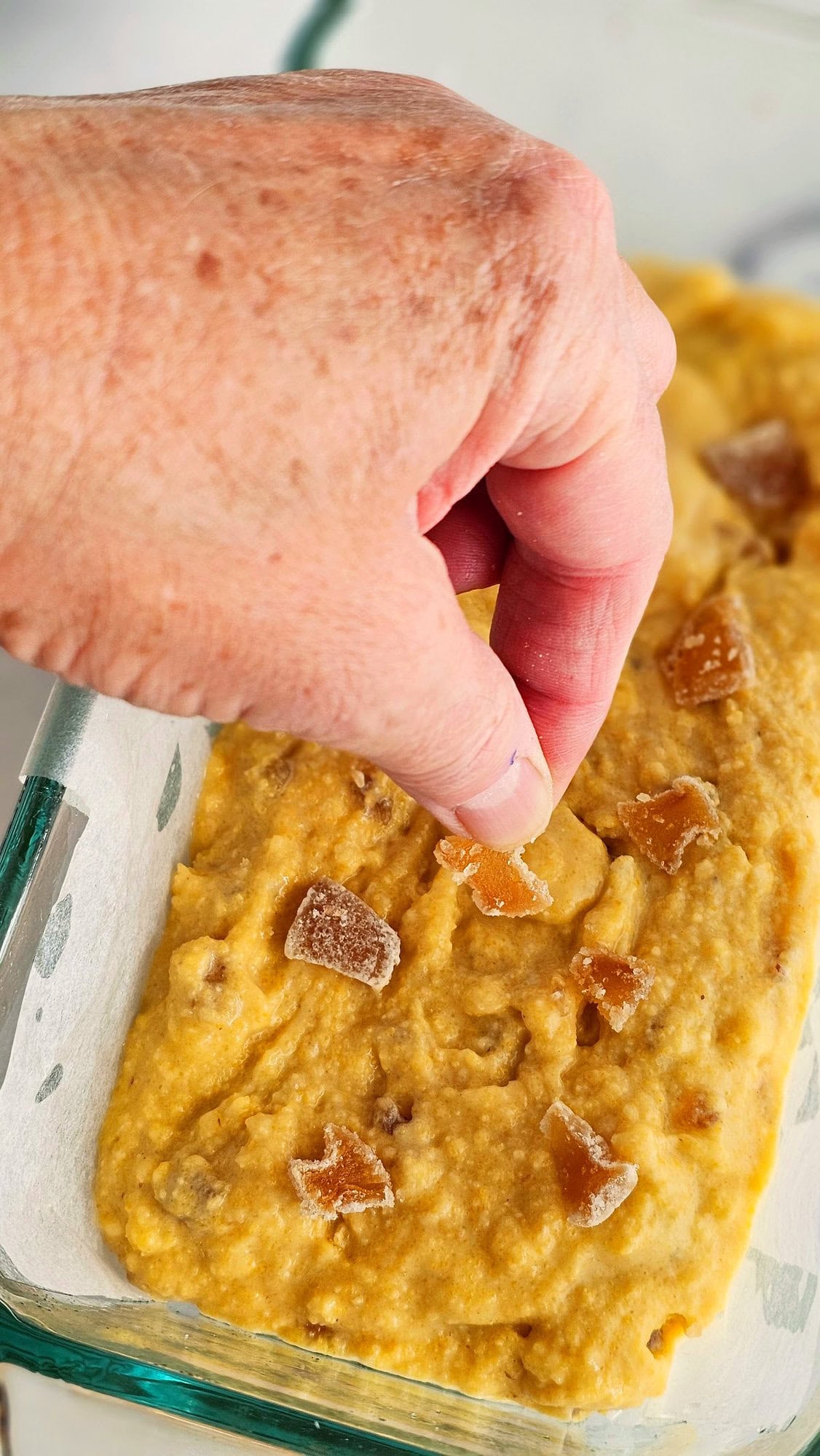 A hand places thin pieces of candied ginger atop batter in a glass loaf pan lined with parchment paper